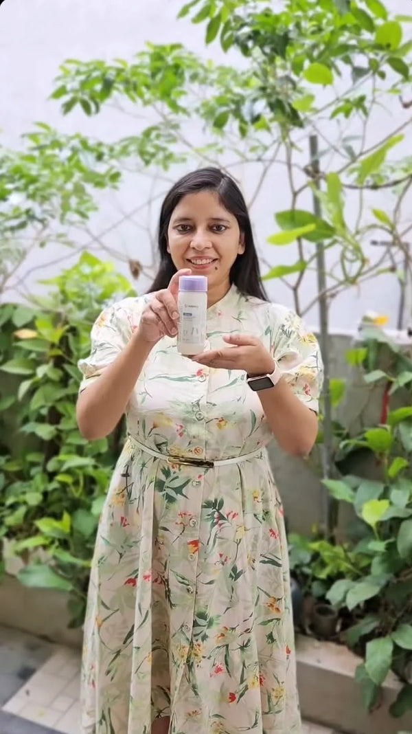 A woman wearing a light beige floral dress holds up a small white bottle with a purple cap in front of a green garden background.