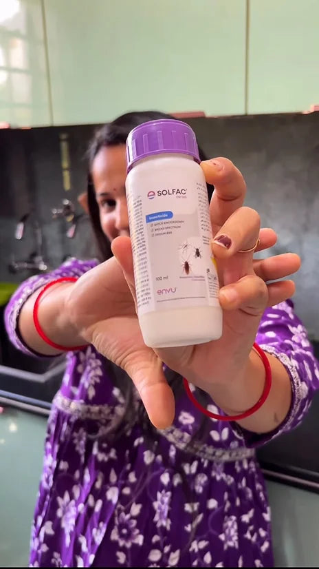 A person in a purple floral dress holds up a white bottle of SOLFAC insecticide with a purple cap in front of a kitchen background.