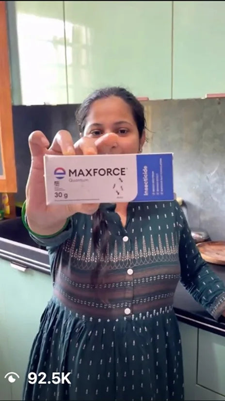 A woman in a patterned green dress holds up a box of MAXFORCE Insecticide in a kitchen setting.